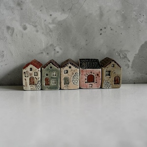 Könnte beinhalten: Ein Set aus fünf Miniatur-Hausfiguren aus Keramik, die in einer Reihe angeordnet sind. Jedes Haus weist einzigartige architektonische Details auf, darunter kleine Fenster, Bogentüren und strukturierte Wände in Creme-, Salbeigrün-, Rosa- und Brauntönen.