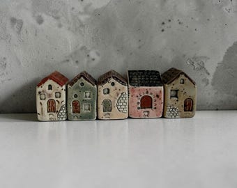 May include: A set of five miniature ceramic houses arranged in a row. Each house features unique hand-painted details, including arched doors, small windows, and textured brick patterns in shades of cream, sage green, terracotta, and dark brown.