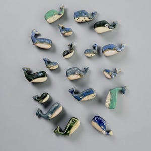 May include: A collection of seventeen small ceramic whale figurines in various shades of blue, green, and cream. Each whale features a smooth, glazed finish with distinct tail and fin shapes, arranged in a scattered pattern on a neutral surface.