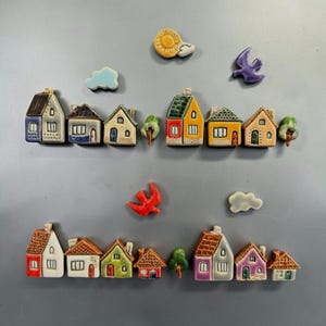 May include: A collection of ceramic magnets featuring miniature houses, trees, clouds, and birds in various colors. The set includes two rows of small, hand-painted structures with detailed roofs, windows, and doors, arranged with decorative sky elements.