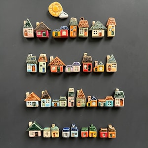 Peut inclure: Une collection de petites figurines de maisons en céramique peintes à la main, disposées en quatre rangées horizontales, présentant diverses formes architecturales, textures de toit et couleurs vives, avec une pièce en céramique soleil et nuage.