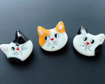 May include: Three ceramic cat head figurines. Each cat head is white with black, orange, and pink accents. The cats have a red heart-shaped nose and whiskers. The figurines are on a dark gray surface.