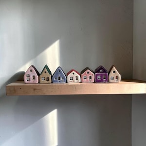 Può includere: Una fila di case in ceramica in miniatura in vari colori pastello, tra cui rosa, verde e viola, è esposta su una mensola di legno. Ogni casa ha un design unico con finestre e porte, creando una scena di villaggio affascinante. Le case sono disposte contro un muro neutro.