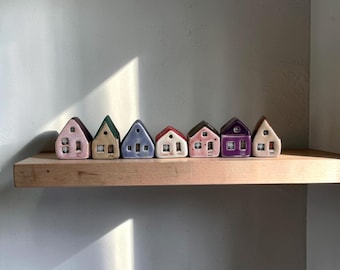 Peut inclure: Une rangée de petites maisons en céramique aux couleurs pastel, dont rose, vert et violet, est exposée sur une étagère en bois. Chaque maison a un design unique avec des fenêtres et des portes, créant une scène de village charmante. Les maisons sont disposées contre un mur neutre.