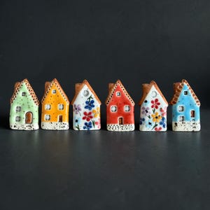 May include: Six miniature ceramic house figurines arranged in a row. Each house features a unique color, including light green, yellow, white with floral patterns, red, white with floral patterns, and blue. All have brown roofs and speckled bases.