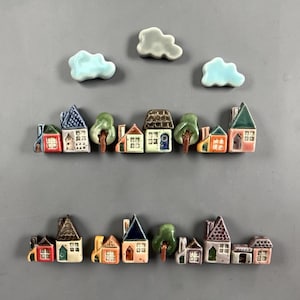 May include: A collection of small ceramic house and tree figurines arranged in two rows, accompanied by three cloud-shaped pieces. The houses feature various roof shapes, colors, and window details, while the trees are green with brown trunks.