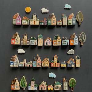 Op de afbeelding: Een verzameling kleurrijke keramische miniatuurhuisjes, bomen, wolken en een zon. De huizen hebben verschillende architecturale stijlen en kleuren, waaronder roze, blauw, geel en groen. De bomen zijn groen en de wolken zijn wit en blauw.
