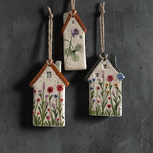 May include: Three ceramic house-shaped ornaments hanging from jute twine. Each piece features a white base with black speckles and hand-painted floral designs. The roofs are textured and coloured in shades of brown or blue, with small windows.