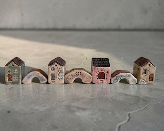 Peut inclure: Une collection de sept structures miniatures en céramique disposées en rangée, comprenant quatre petites maisons aux toits en pente et trois pièces en forme d'arche, avec des émaux pastel variés dans des tons vert, rose, beige et blanc moucheté.