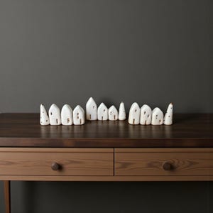 May include: A collection of white ceramic miniature houses with gold accents, arranged in a row on a wooden surface. Each house has a unique shape and design, with small windows and doors. The background is a solid gray wall, creating a minimalist aesthetic.
