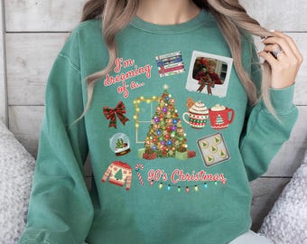 May include: A teal green sweatshirt with a festive 90's Christmas design. The graphic includes a Christmas tree, a snow globe, a sweater, a bow, a photo, mugs, cookies, and the text "I'm dreaming of... 90's Christmas."