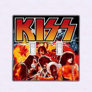 May include: A double light switch cover featuring a colorful image of the rock band KISS. The band's name is written in large, bold letters across the top of the image. The band members are shown in a fiery, energetic pose.