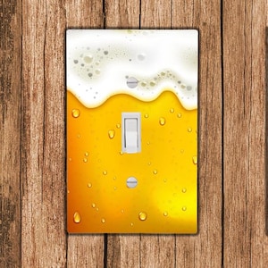 Op de afbeelding: Een lichtschakelaar-afdekplaat met een afbeelding van een glas bier met schuim erop. Het bier is geel en het schuim is wit.