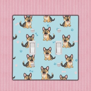 Op de afbeelding: Een lichtblauwe dubbele lichtschakelaar met een herhalend patroon van cartoon Duitse herdershonden, pootafdrukken en hondenbotten. De honden zijn zwart, bruin en wit. De afdekking heeft twee witte schakelaars.