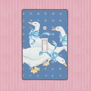May include: A decorative light switch cover featuring a blue background with a pattern of white flowers. The cover has a watercolor illustration of three white geese with blue bows around their necks. The switch is white.