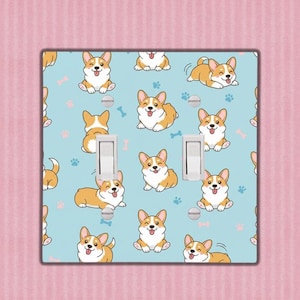 Op de afbeelding: Een lichtblauwe dubbele lichtschakelaar met een herhalend patroon van cartoon corgi honden, pootafdrukken en botten. De corgi's staan in verschillende poses, met witte en bruine vacht. De afdekking heeft twee witte lichtschakelaars.