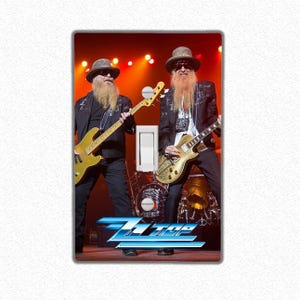 May include: A light switch cover featuring a photograph of two musicians playing guitars. The musicians are wearing hats and have long beards. The band's name "ZZ Top" is printed in blue on the bottom of the cover.