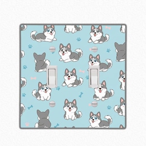 Op de afbeelding: Een lichtblauwe dubbele lichtschakelaar cover met een herhaald patroon van cartoon Siberische Husky honden in grijs en wit. Het ontwerp bevat pootafdrukken, botten en hondenbakken.