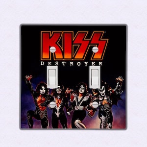 以下が含まれることがあります： 「デストロイヤー」のアルバムカバーアートが描かれたKISSのバンドをフィーチャーしたライトスイッチプレート。画像には、4人のバンドメンバーが特徴的なメイクとコスチュームで写っています。