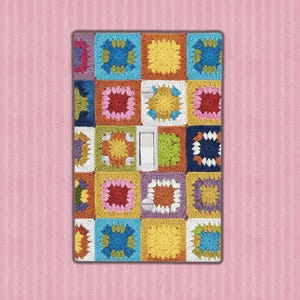 May include: A colorful light switch cover featuring a patchwork design. The cover is adorned with a grid of crocheted squares in various colors, including yellow, blue, orange, and pink. A white toggle switch is centered on the cover.
