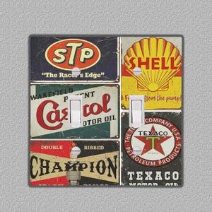 Puede incluir: Una cubierta decorativa para interruptores de luz con logotipos publicitarios vintage. El diseño incluye logotipos de STP, Shell, Castrol Motor Oil, Champion y Texaco. La cubierta tiene dos interruptores.