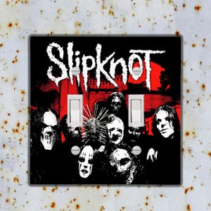 May include: A black and white light switch cover featuring a band logo for Slipknot. The logo is in white with a red background. The band members are wearing masks and are in the background.