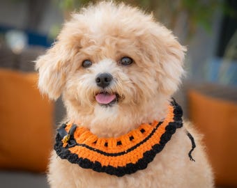 May include: A small, fluffy dog with light brown fur wearing a handmade orange and black crochet collar. The collar has a scalloped edge and a small bow. The dog is looking directly at the camera with its tongue out.