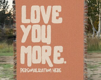 May include: A woven blanket in a terracotta color, featuring the words "LOVE YOU MORE" in large, off-white letters. The phrase "PERSONALIZATION HERE" is below. The blanket has a fringed edge.