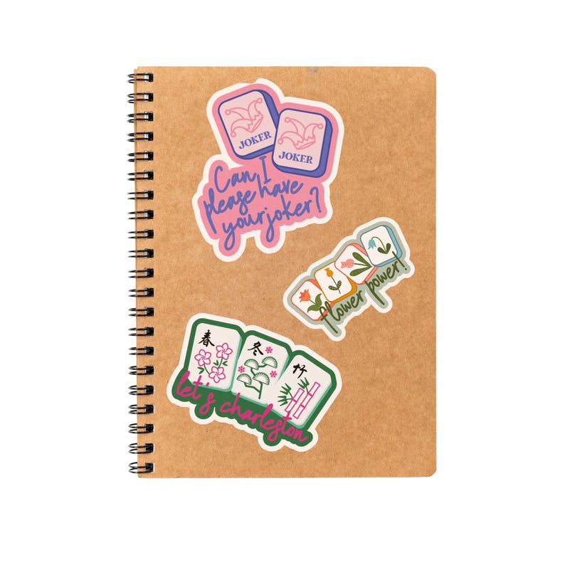 K&ouml;nnte beinhalten: Ein Spiralnotizbuch mit einem Kraftpapier-Einband, verziert mit bunten Aufklebern. Die Aufkleber zeigen S&auml;tze wie "Can I please have your joker?" und "Flower power!" zusammen mit Mahjong-Kachel-Designs.
