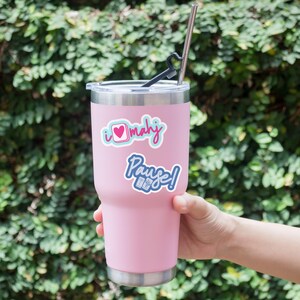 K&ouml;nnte beinhalten: Ein rosa isolierter Becher mit einem silbernen Strohhalm und einem schwarzen Strohhalmhalter. Der Becher hat zwei Aufkleber mit der Aufschrift "i ❤️ mahj" und "Pause!". Der Becher wird von einer Person vor einem gr&uuml;nen, belaubten Hintergrund gehalten.