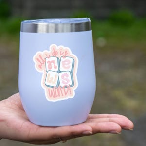 K&ouml;nnte beinhalten: Ein hellblaues, isoliertes Weinglas mit silbernem Rand, versehen mit einem Aufkleber mit der Aufschrift "all I do is NEWS WINE!" in Pastellfarben. Das Glas wird in einer Hand gehalten, mit einem verschwommenen gr&uuml;nen Hintergrund.