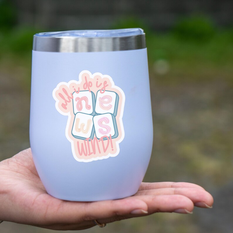 K&ouml;nnte beinhalten: Ein hellblaues, isoliertes Weinglas mit silbernem Rand, versehen mit einem Aufkleber mit der Aufschrift "all I do is NEWS WINE!" in Pastellfarben. Das Glas wird in einer Hand gehalten, mit einem verschwommenen gr&uuml;nen Hintergrund.