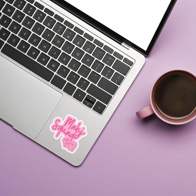 K&ouml;nnte beinhalten: Ein Laptop mit schwarzer Tastatur und eine rosa Kaffeetasse auf einer lila Oberfl&auml;che. Ein rosa Aufkleber mit den Worten "Mahj Squad" und zwei rosa Kacheln befindet sich auf dem Laptop.