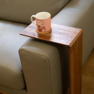 May include: A wooden side table with a rectangular top and a curved side. The table is attached to the arm of a grey sofa and has a pink mug with a floral design on it.
