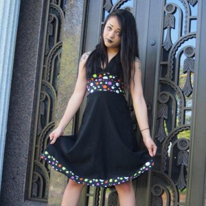 K&ouml;nnte beinhalten: Eine Frau in einem schwarzen Kleid mit buntem Tupfenmuster und wei&szlig;em Saum. Sie steht vor einer gro&szlig;en, verzierten T&uuml;r.
