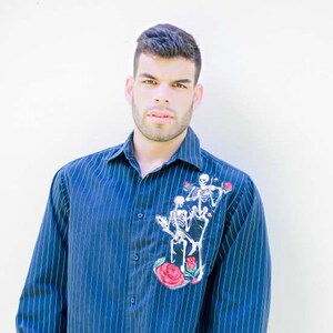 May include: A man wearing a blue striped button-down shirt with a skeleton design and red roses. The shirt has a white background with thin blue stripes. The man is wearing blue jeans and has short brown hair.