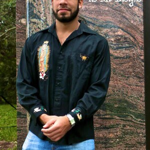 May include: A black long-sleeve button-down shirt with embroidered details. The shirt features a religious image and a crown on the left chest. The shirt is worn by a person with a beard and a casual expression.