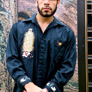 May include: A man wearing a dark blue long-sleeved shirt with embroidered details. The shirt features a religious image of the Virgin Mary and a crown. The man is standing in front of a textured wall.