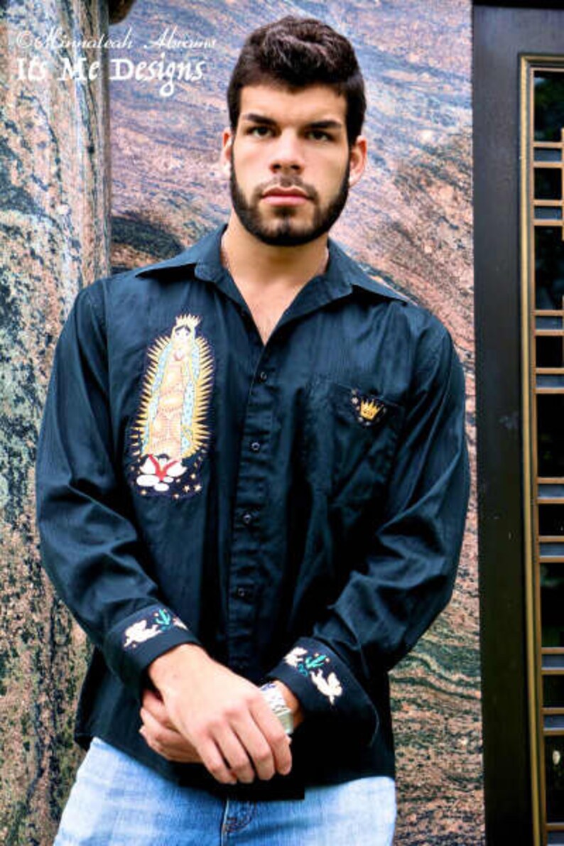 May include: A man wearing a dark blue long-sleeved shirt with embroidered details. The shirt features a religious image of the Virgin Mary and a crown. The man is standing in front of a textured wall.