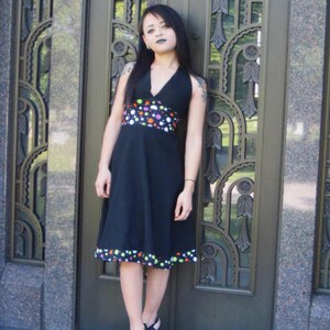 K&ouml;nnte beinhalten: Eine Frau tr&auml;gt ein schwarzes Halfterkleid mit einem bunten Tupfenband um die Taille. Das Kleid hat einen bunten Tupfenbesatz am Saum. Sie tr&auml;gt schwarze Sandaletten mit Absatz.