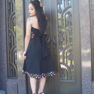 K&ouml;nnte beinhalten: Eine Frau in einem schwarzen Kleid mit wei&szlig;en Punkten und einem bunten Saum. Das Kleid hat einen tr&auml;gerlosen Ausschnitt und eine Schleife am R&uuml;cken. Sie steht vor einer dekorativen Metallt&uuml;r.
