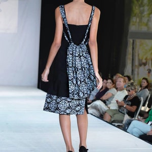 May include: A model walks the runway wearing a black and white patterned dress with a black bodice and a white skirt. The dress has a unique design with a black overlay and a white skirt with a floral pattern. The model is wearing black heels.