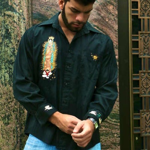 May include: A man wearing a dark green button-down shirt with a white embroidered image of the Virgin Mary. The shirt has a white embroidered design on the cuffs. The man is wearing blue jeans.