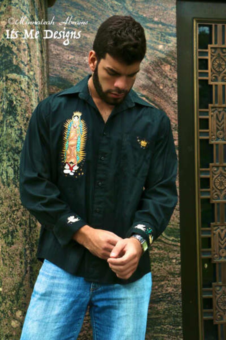 May include: A man wearing a dark green button-down shirt with a white embroidered image of the Virgin Mary. The shirt has a white embroidered design on the cuffs. The man is wearing blue jeans.