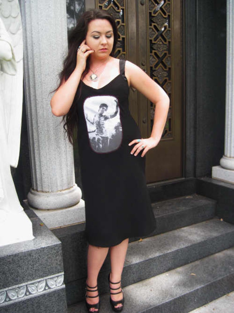 Puede incluir: Una mujer lleva un vestido negro sin mangas con un estampado gr&aacute;fico de una persona con una camisa blanca. El vestido tiene un escote redondo y una silueta ajustada. Lleva tacones negros y tiene el pelo recogido hacia atr&aacute;s.