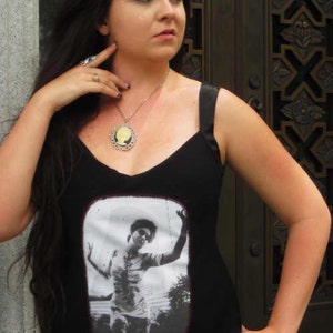 Puede incluir: Una mujer con el pelo largo y oscuro lleva un top negro de tirantes con una imagen de camafeo de una mujer en la parte delantera. Lleva un collar de plata con un colgante de camafeo. Tambi&eacute;n lleva un anillo de plata en la mano izquierda y un anillo turquesa en la mano derecha.
