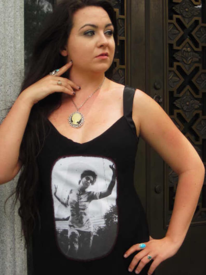 Puede incluir: Una mujer con el pelo largo y oscuro lleva un top negro de tirantes con una imagen de camafeo de una mujer en la parte delantera. Lleva un collar de plata con un colgante de camafeo. Tambi&eacute;n lleva un anillo de plata en la mano izquierda y un anillo turquesa en la mano derecha.