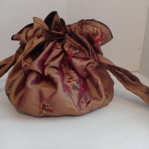 Könnte beinhalten: Eine braune und goldene Kordeltasche mit Blumenmuster. Die Tasche ist aus seidenartigem Stoff und hat einen langen, dünnen Riemen.