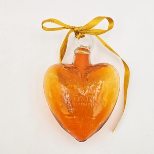 May include: A heart-shaped glass ornament with a gold ribbon. The ornament is amber in color and has a textured surface. The ornament has the text "La Venta del Paraiso" on it.
