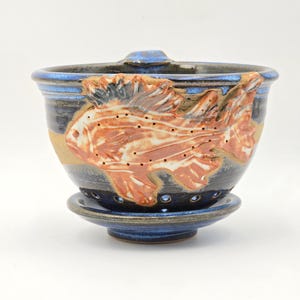 May include: A ceramic tea strainer with a fish design. The strainer is blue with a brown and white fish relief. The strainer sits on a matching blue saucer. The rim of the strainer has a blue stripe.